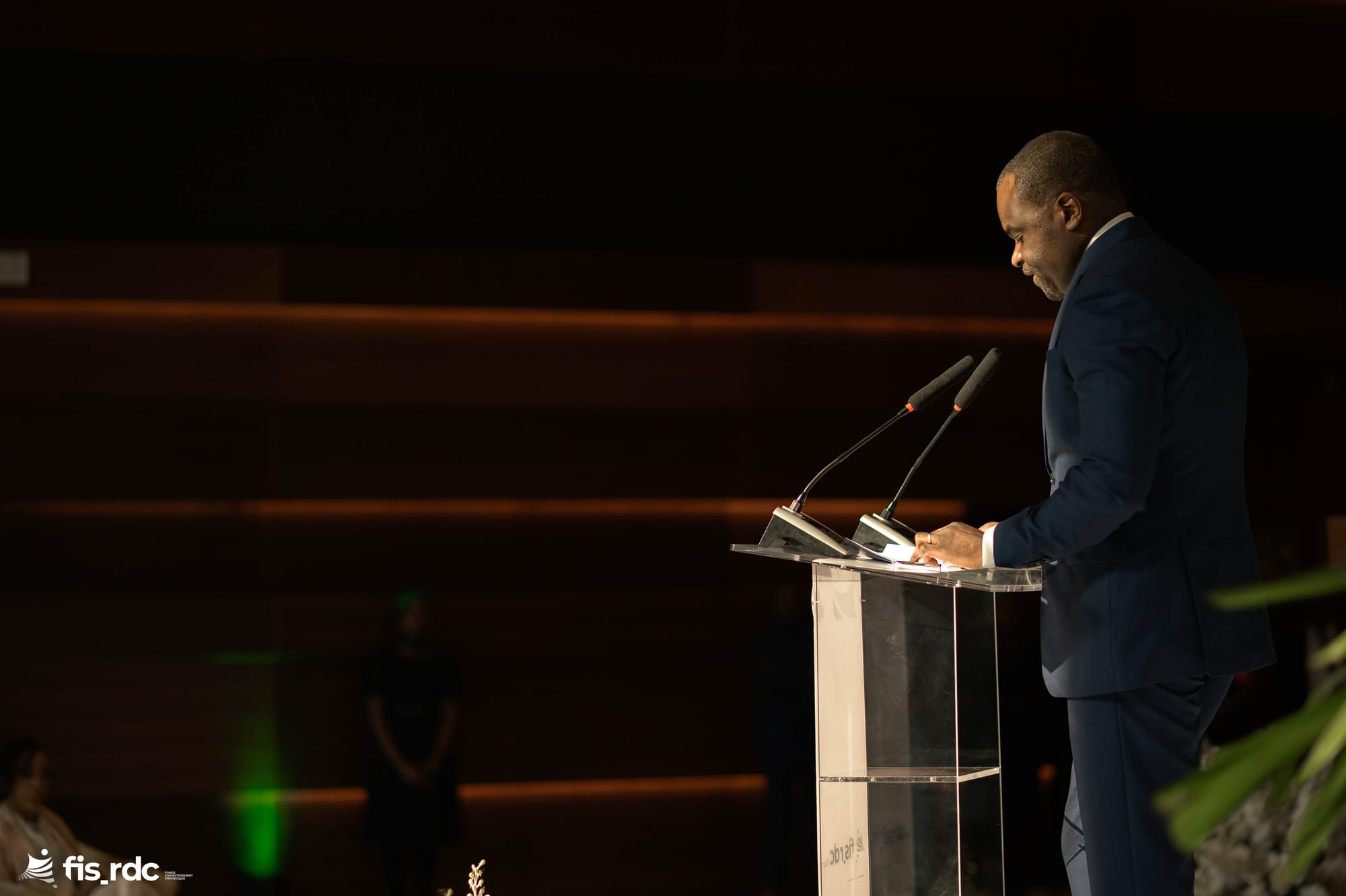 Émile OSUMBA au pupitre, dans la lumière de la scène.