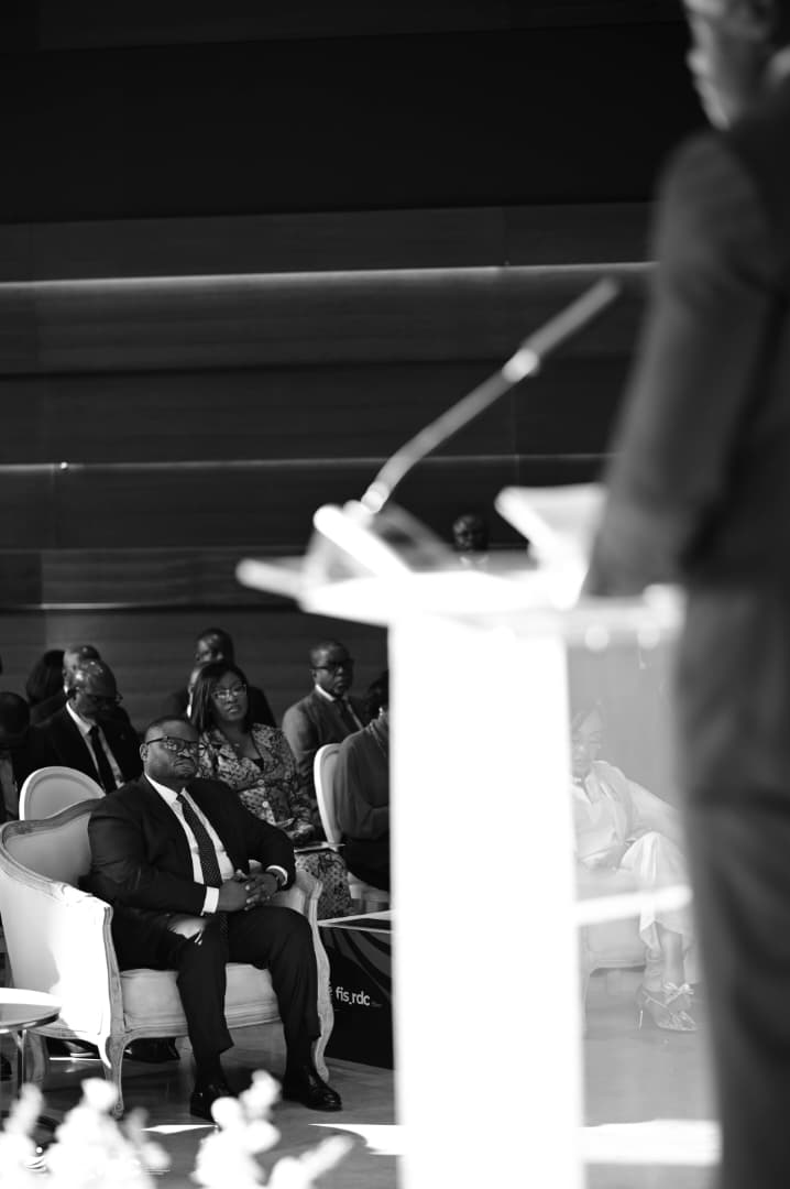 Les officiels écoutent une allocution, vus depuis l’arrière du pupitre (noir et blanc).